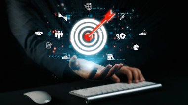 Bir işadamı, karanlık bir arkaplana karşı strateji, teknoloji ve takım çalışmasını temsil eden çeşitli simgelerle çevrili dijital hedef grafiğine uzanıyor. Dörtgen