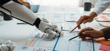 Bir robot kol ve insan eli modern bir ofiste iş verilerini ve grafiklerini analiz ederek teknoloji ve insan uzmanlığının kaynaşmasını gösteriyor. Noogenesis