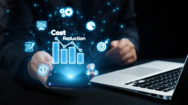 Bir işletme profesyoneli, maliyetlerin azaltılması ile ilgili dijital grafik ve simgeleri gösteren bir akıllı telefon sergileyerek, modern araçları ve finans alanında stratejik planlamayı vurguluyor. Dörtgen
