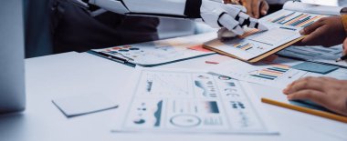 Fütüristik bir ofis sahnesinde çeşitli veri ve çizelgeleri analiz eden, takım çalışmalarını ve yeniliklerini geliştiren bir grup uzmana yardımcı olan robotik bir kol sergileniyor.