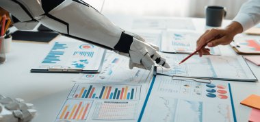 Bu resim, insan eliyle çalışan bir robot kolu yakalıyor. Çeşitli iş haritaları ve grafikler üzerinde modern otomatikman sergiliyor. Noogenesis