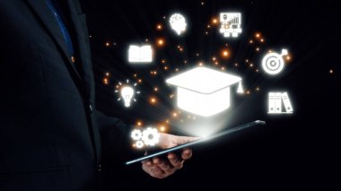 Bir işadamı, teknoloji ve öğrenmenin kesişimlerini gösteren çeşitli eğitim sembolleriyle aydınlatılmış bir tablet tutuyor. Görüntü yenilik ve bilgiyi temsil ediyor. Dörtgen