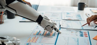 Gelecekçi bir robot kolu, bir ofis masasında insan eliyle iş raporları üzerinde işbirliği yaparak teknoloji ve insan çabasının veri analizlerindeki sinerjisini gösteriyor. Noogenesis