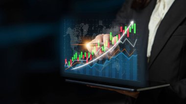 A business professional examines a vibrant digital graph on a laptop, showcasing financial growth and market trends in a modern office setting at night. Impute