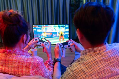 Kontrollü joystickli birkaç oyuncu arka tarafta televizyon ekranında dövüş oyunu oynuyor, modern rahat evinde neon ışıklı renkli oturma odasında koltukta oturuyor. İnfobahn.