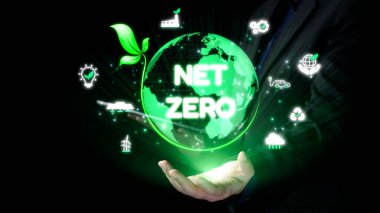 Yeşil parıldayan bir küreyi tutan bir el, sürdürülebilirlik, teknoloji ve çevresel sorumluluk sembolleriyle çevrili net sıfır hedef kavramını temsil eder. Dörtgen