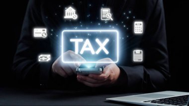 Parlak vergi sembolleri ve finansal planlama ve yönetim simgelerini gösteren bir akıllı telefon kullanan bir kişi. Karanlık arka plan dijital odağı arttırır. Dörtgen