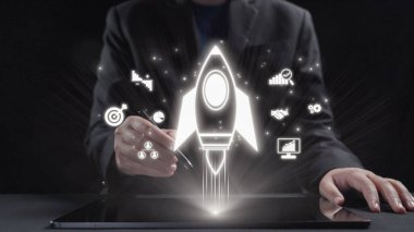 Bir iş adamı, modern çalışma alanındaki büyüme, teknoloji ve yaratıcılığı vurgulayan yenilikçi projelerin başlatılmasını gösteren bir roket ikonunu görselleştirmek için bir tablet kullanır. Dörtgen