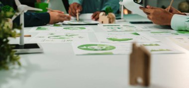 Bir grup profesyonel, sürdürülebilir projeler üzerinde yoğunlaşarak çağdaş bir ofiste işbirliği yapıyor. Belgeler, çizelgeler ve planlar yayılmış, yenilik ve takım çalışması sergileniyor. SACTR