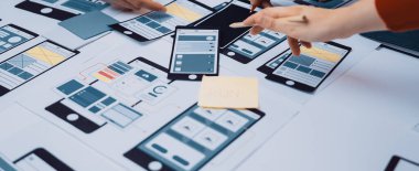 Bir masanın üzerinde, takım çalışması ve yaratıcılığı vurgulayan, çizimler, prototipler ve proje notlarıyla ilgilenen, işbirlikçi bir uygulama tasarım sürecini gösteren dinamik bir çalışma alanı. SACTR