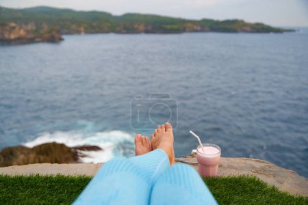 Photo for A young woman with bare feet sits on the edge of a cliff above the ocean. A first-person photograph - Royalty Free Image