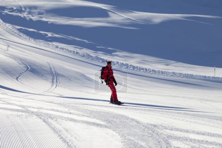 Téléchargez les photos : Snowboarder en descente rouge sur piste de ski enneigée et ensoleillée préparée par la toiletteuse lors d'une soirée d'hiver ensoleillée - en image libre de droit