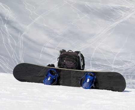 Photo for Snowboard and black backpack on snow in high winter mountains and snowy off-piste slope with traces of skis and snowboards at background - Royalty Free Image