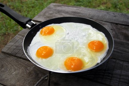 Téléchargez les photos : Old wooden table and frying pan with fried eggs on camping gas stove. Outdoor cooking. - en image libre de droit