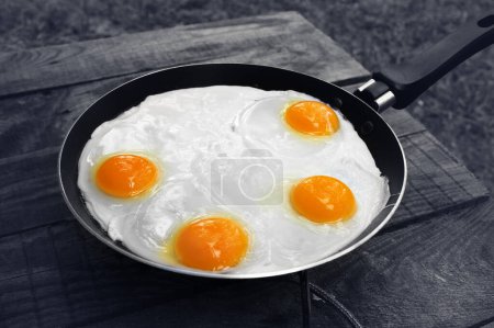 Photo for Black and white old wooden table and frying pan with fried eggs on camping gas stove. Outdoor cooking. Selective color effect. - Royalty Free Image