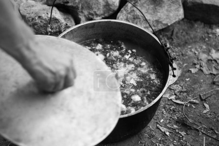 Photo for Freshly cooked hot soup in old big sooty cauldron on camp fire and man hand with pot cover. Outdoor camping cooking. Selective focus. Black and white toned image. - Royalty Free Image