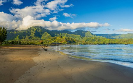 Téléchargez les photos : Panorama de la plage de sable fin de la baie de Hanalei avec jetée et montagnes Na Pali entourées de nuages ensoleillés - en image libre de droit