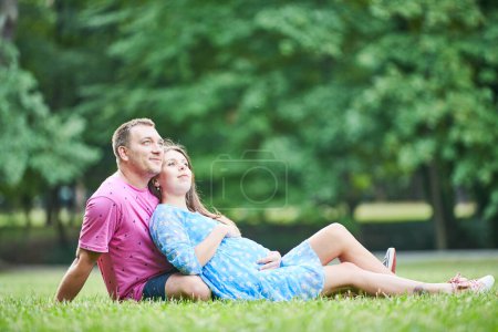 Téléchargez les photos : Couple, y compris jeune homme et femme enceinte assis sur l'herbe dans le parc. Concept de famille, de grossesse et de maternité - en image libre de droit