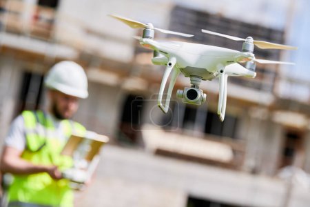 Téléchargez les photos : Un travailleur de la construction pilote un drone sur le chantier. Surveillance vidéo ou inspection de la sécurité industrielle ou tournage aérien par un ingénieur spécialiste en architecture avec drone volant. Focus sur drone - en image libre de droit