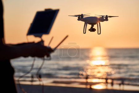 Foto de Hombre operando dron al atardecer. Drone volando sobre un mar. Concéntrate en drone - Imagen libre de derechos
