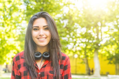 Photo for Beautiful multiracial woman portrait with headphones around the neck - Happy girl in Italy during summer looking at camera and smiling, green trees and backlight on background - Royalty Free Image