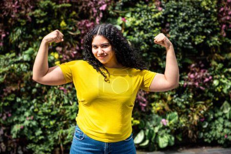 Téléchargez les photos : Young multiracial woman flexing muscles and looking at camera. Multiracial curly girl smiling and showing arms in a playful way, showing confidence and happiness. Lifestyle and diversity concepts - en image libre de droit
