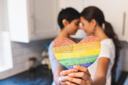 Photo for Authentic shot of happy lesbian couple holding a heart shaped rainbow flag and hugging - lesbian couple at home enjoying life together - Royalty Free Image