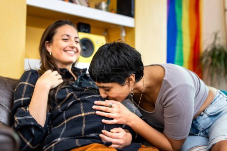 Téléchargez les photos : Authentic shot of happy married homosexual female gay couple with woman kissing tummy of pregnant partner or friend, rainbow pride flag on background - lesbian couple at home enjoying life together - en image libre de droit