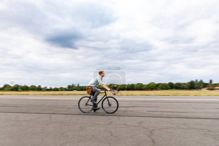 Téléchargez les photos : Homme faisant du vélo dans un parc de la ville - Homme souriant allant sur un vélo à engrenages fixes pour faire des livraisons ou des trajets domicile-travail - effet de mouvement avec technique de panoramique - en image libre de droit