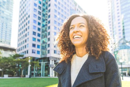 Foto de Retrato exitoso de mujer de negocios en la ciudad - Mujer negra sonriente con rizos disfrutando de un día soleado - Vestido casual inteligente - Conceptos de estilo de vida y felicidad - Imagen libre de derechos