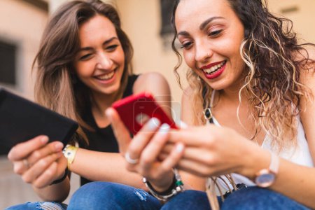 Foto de Millennials niñas felices usando teléfonos móviles y riendo, disfrutando del tiempo juntos en la ciudad en un día de verano - Estilo de vida, conceptos de amistad de personas felices utilizando dispositivos tecnológicos para redes sociales - Imagen libre de derechos