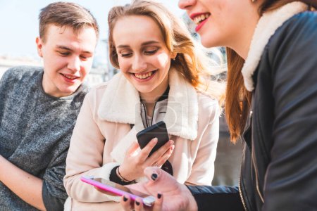 Foto de Feliz grupo de amigos usando smartphone y riéndose - Tres jóvenes, un hombre y dos mujeres, relajándose y divirtiéndose, sonriendo y hablando juntos - Conceptos de amistad y estilo de vida - Imagen libre de derechos