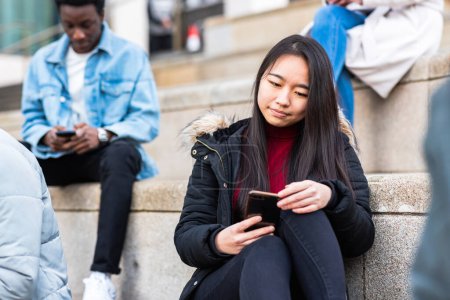 Foto de Multiracial millennial personas que utilizan teléfonos inteligentes - Mujer asiática en primer plano y la celebración de un teléfono inteligente móvil, entornos urbanos- Estudiantes universitarios o amigos sentados y teniendo un descanso en la universidad - Imagen libre de derechos
