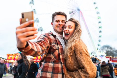 Foto de Pareja feliz divirtiéndose en el parque de atracciones de Londres - pareja joven enamorada de tomar una selfie y disfrutar del tiempo en la feria con montaña rusa en el fondo - Estilo de vida feliz y conceptos de amor - Imagen libre de derechos