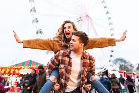 Téléchargez les photos : Un couple heureux qui s'amuse au parc d'attractions de Londres - Portrait d'un jeune couple amoureux qui s'amuse à la fête foraine avec des montagnes russes en arrière-plan - Happy lifestyle and love concepts - en image libre de droit