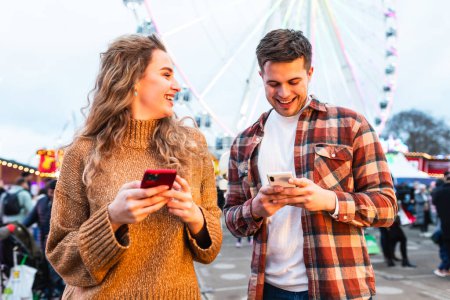 Téléchargez les photos : Couple heureux au parc d'attractions en regardant le téléphone portable et en riant - jeune couple s'amuser ensemble lors d'une soirée à la foire d'amusement - concepts d'amour et de bonheur - en image libre de droit