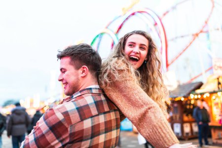 Photo for Happy couple having fun at amusement park in London - Portrait of young couple in love enjoying time at funfair with rollercoaster on background - Happy lifestyle and love concepts - Royalty Free Image