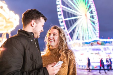 Foto de Pareja feliz en el parque de atracciones mirando el teléfono móvil y riendo - pareja joven divirtiéndose juntos en una noche en la feria de la diversión - conceptos de amor y felicidad - Imagen libre de derechos