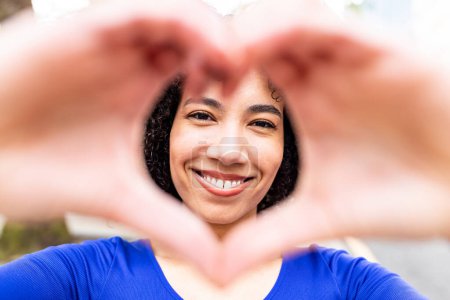 Photo for Happy young woman gesturing heart shape with hands, looking through making a nice smile to the camera - Portrait of beautiful black curly woman showing love sign - Royalty Free Image