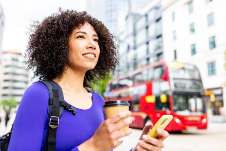 Foto de Mujer feliz con el pelo rizado y aspecto multiétnico en la ciudad - Estilo de vida y transporte en Birmingham - Mujer negra con teléfono inteligente y taza de café conmutando - Imagen libre de derechos