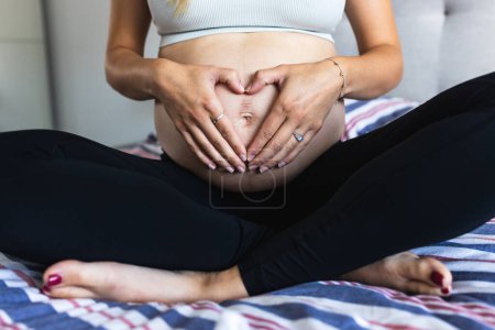 Foto de Mujer embarazada haciendo forma de corazón con las manos en el bulto mientras está sentada en la cama - Primer plano plano de la barriga desnuda durante el embarazo - Un solo padre femenino - Imagen libre de derechos