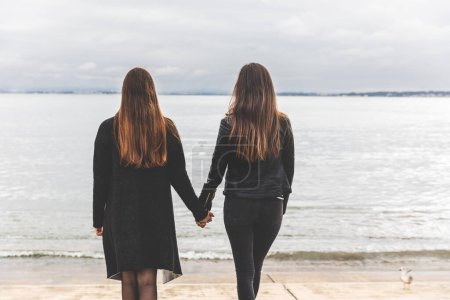 Téléchargez les photos : Vue arrière de deux femmes se tenant la main, regardant au loin un lac ou une rivière tranquille - Deux femmes couple ou amis se tenant la main au bord du lac - en image libre de droit