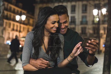 Téléchargez les photos : Homme et femme souriants profitant du contenu du téléphone ensemble dans un cadre de soirée en ville - Heureux couple multiracial partageant un moment avec smartphone la nuit - en image libre de droit