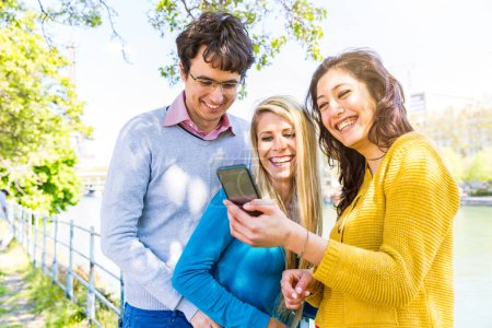Téléchargez les photos : Trois amis joyeux profitant du contenu des médias sociaux sur un téléphone dans un parc ensoleillé de Paris au bord de la Seine et de la tour Eiffel - Amis riant et partageant du contenu sur smartphone en plein air - en image libre de droit