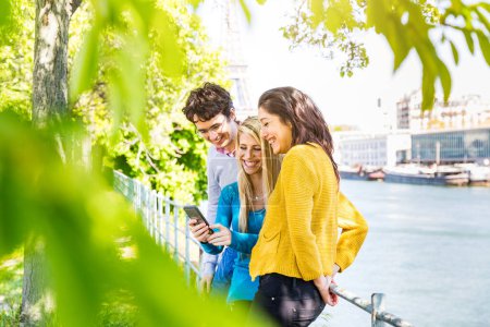 Foto de Tres alegres amigos disfrutando de contenido de redes sociales en un teléfono en un parque soleado en París junto al río Sena y la torre Eiffel - Amigos riendo y compartiendo contenido en teléfonos inteligentes al aire libre - Imagen libre de derechos