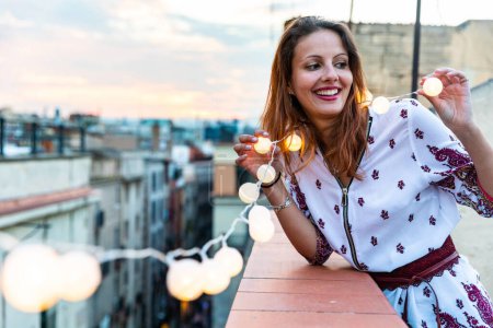 Foto de Feliz retrato de mujer sonriente en una azotea de Barcelona - Joven mujer caucásica con un vestido de flores y sosteniendo luces de hadas - Felicidad y alegría, espacio de copia a la izquierda - Imagen libre de derechos