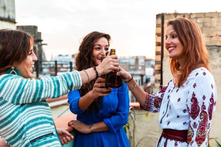 Foto de Mujeres felices disfrutando de una cerveza en una azotea de Barcelona - Tres chicas bebiendo una cerveza y divirtiéndose durante una fiesta al atardecer en una azotea con barrio gótico de fondo en Barcelona - Imagen libre de derechos