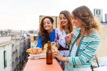 Téléchargez les photos : Femmes heureuses profitant d'un dîner sur un toit à Barcelone - Trois jeunes femmes buvant de la bière et mangeant de la pizza s'amusant lors d'une fête au coucher du soleil sur un toit avec quartier gothique en arrière-plan à Barcelone - en image libre de droit
