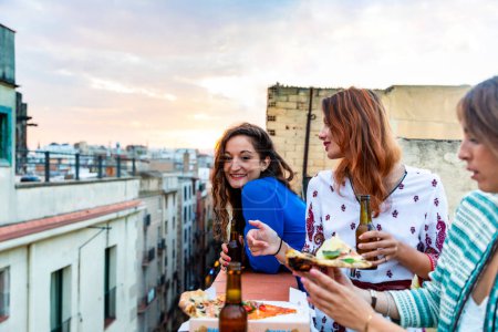 Foto de Mujeres felices disfrutando de una cena en una azotea de Barcelona - Tres mujeres jóvenes bebiendo cerveza y comiendo pizza divirtiéndose durante una fiesta al atardecer en una azotea con barrio gótico de fondo en Barcelona - Imagen libre de derechos