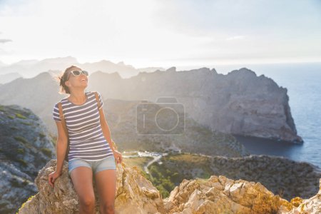 Téléchargez les photos : Joyeuse voyageuse aux lunettes de soleil assise au bord de la mer, admirant un paysage côtier magnifique - Femme souriante jouissant d'une vue panoramique sur la côte - en image libre de droit